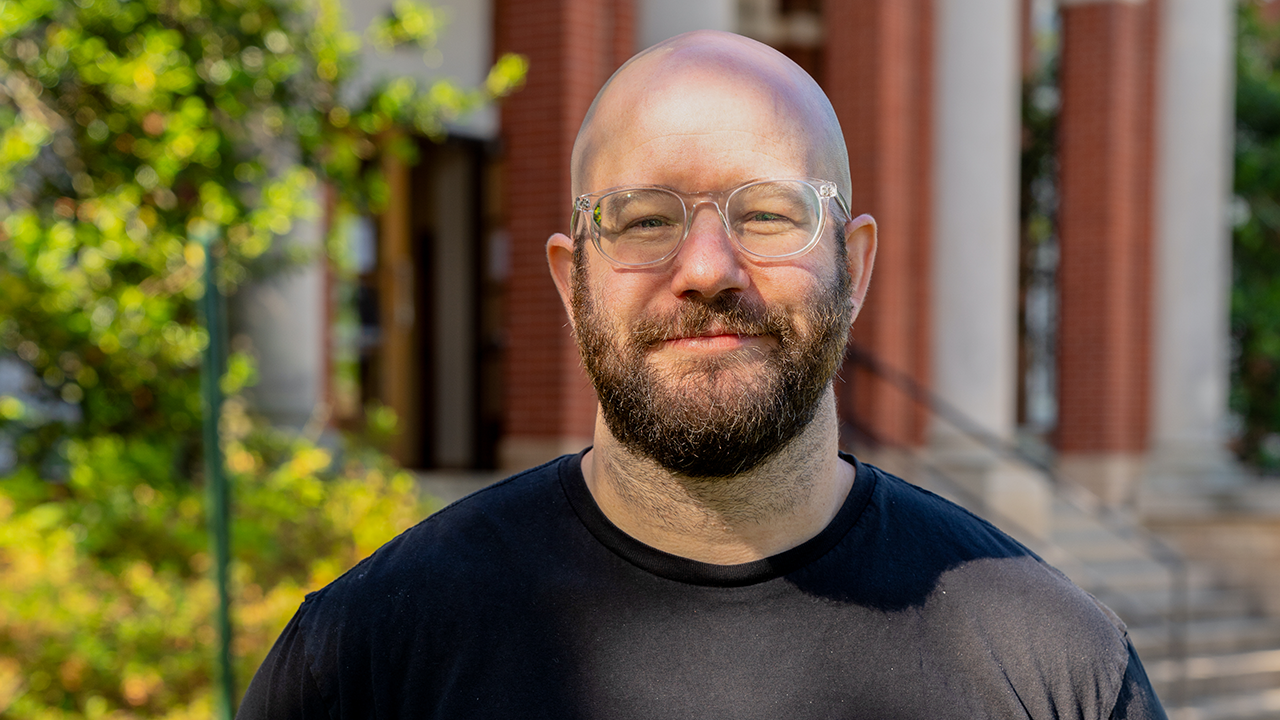 Bald man wearing glasses stands in front of brick building and bushes.