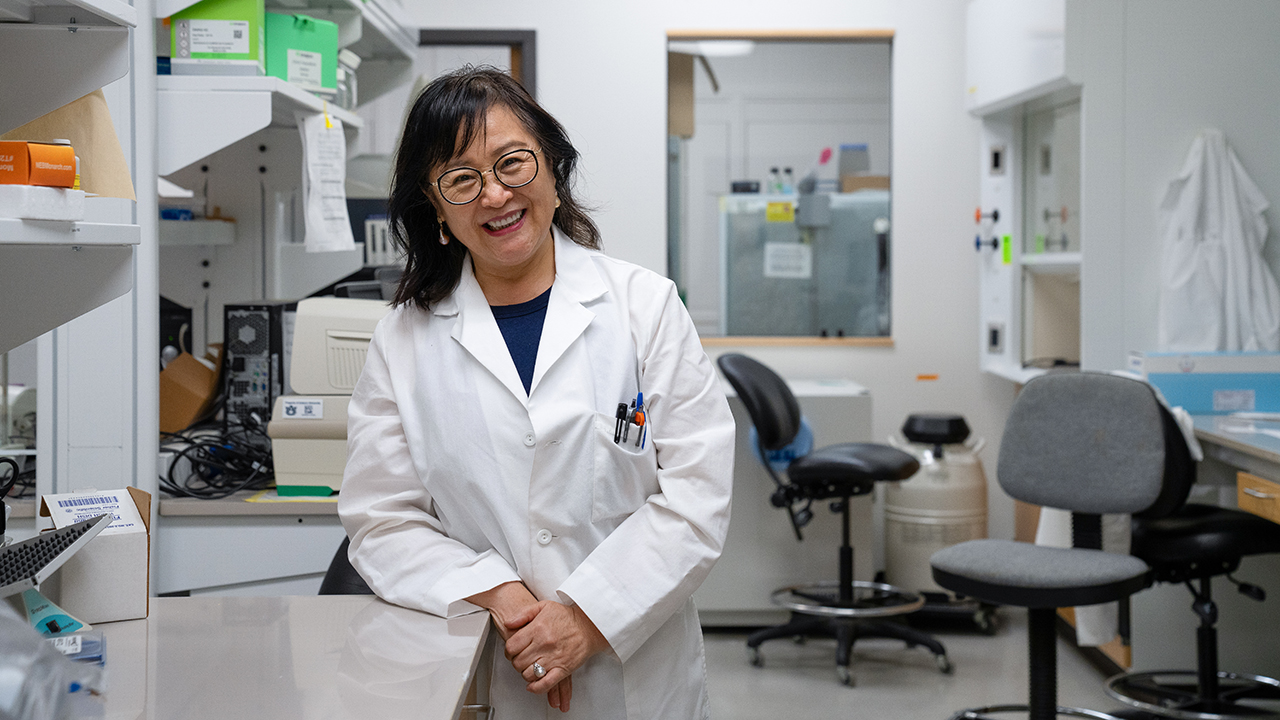 A Chinese woman wears a lab coat and smiles as she leans against a counter in a lab