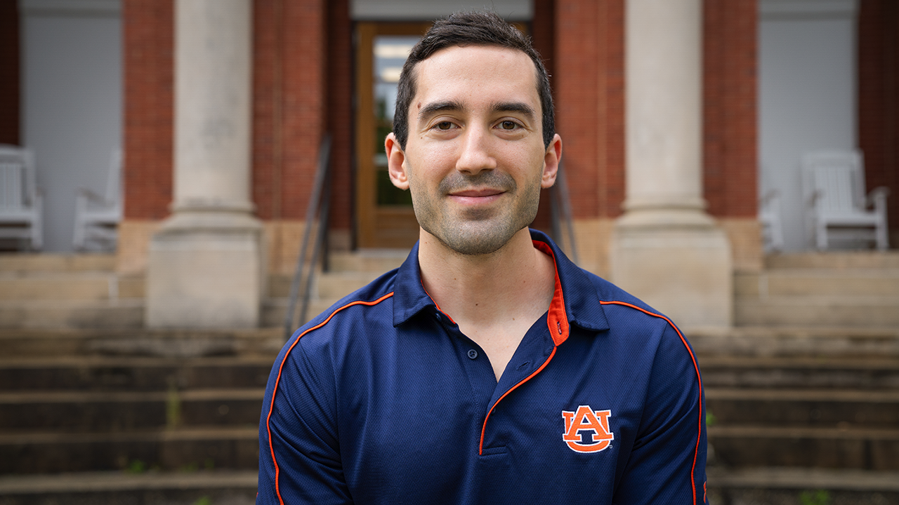 Man wears navy blue polo and smiles in front of brick college building