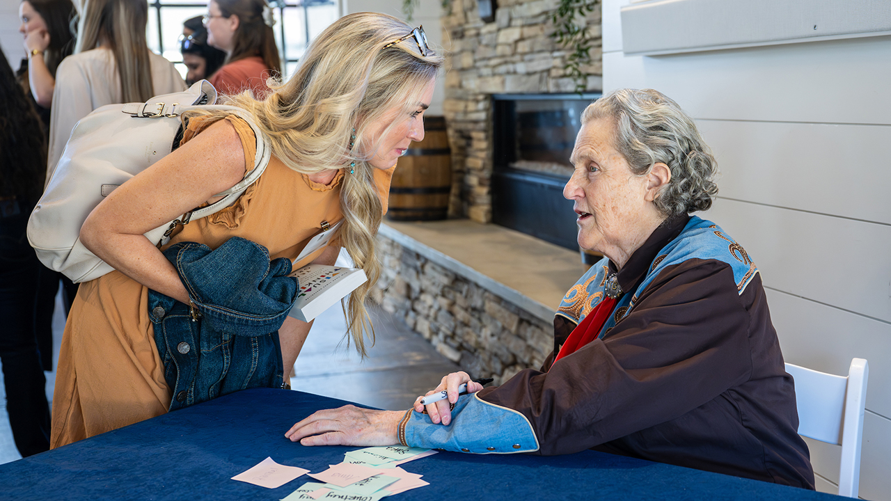 A woman with long blond hair in a pale orange dress bends to speak to Temple Grandin, seated on the right at a table with a blue cloth