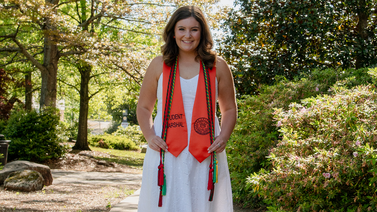 a young woman wears a white dress with graduation tassels and an orange stole that says Student Marshal