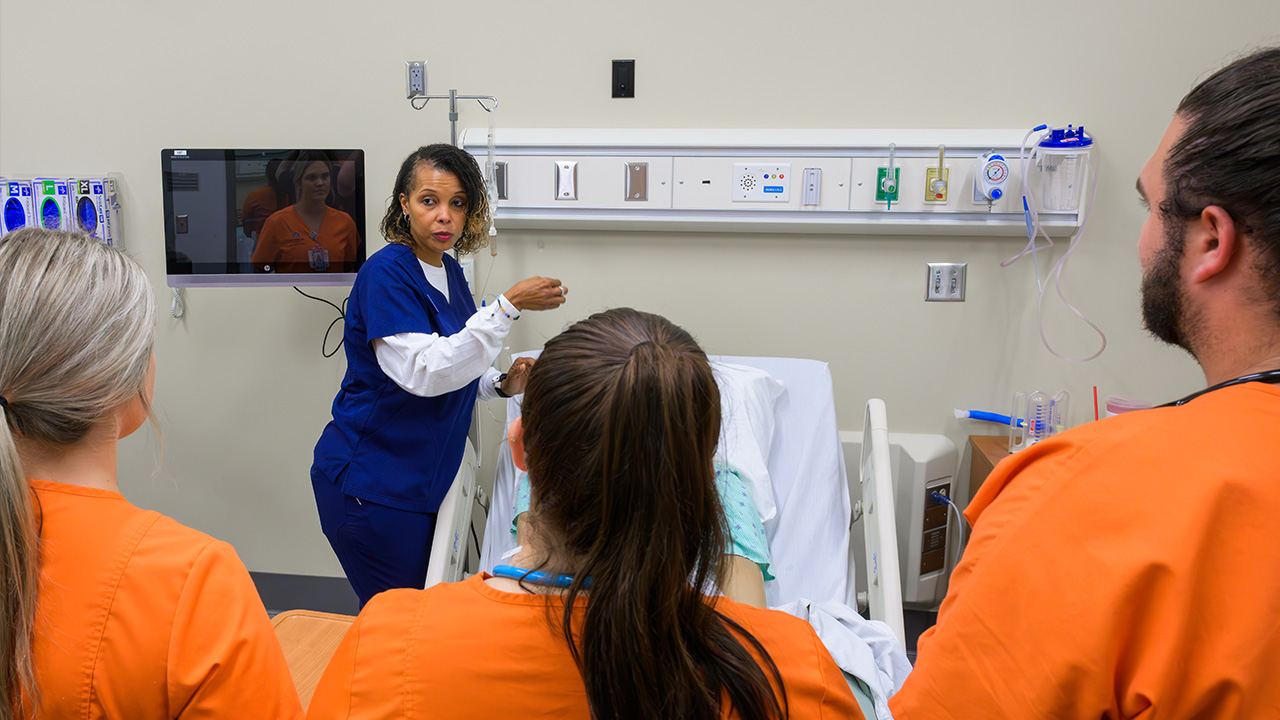 Image of students standing in front of a bed wearing orange scrubs while an instructor talks to them from the head of the bed.