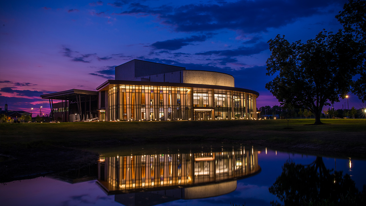 A dusk photograph of the Gogue Performing Arts Center with the interior lights reflecting against a pond