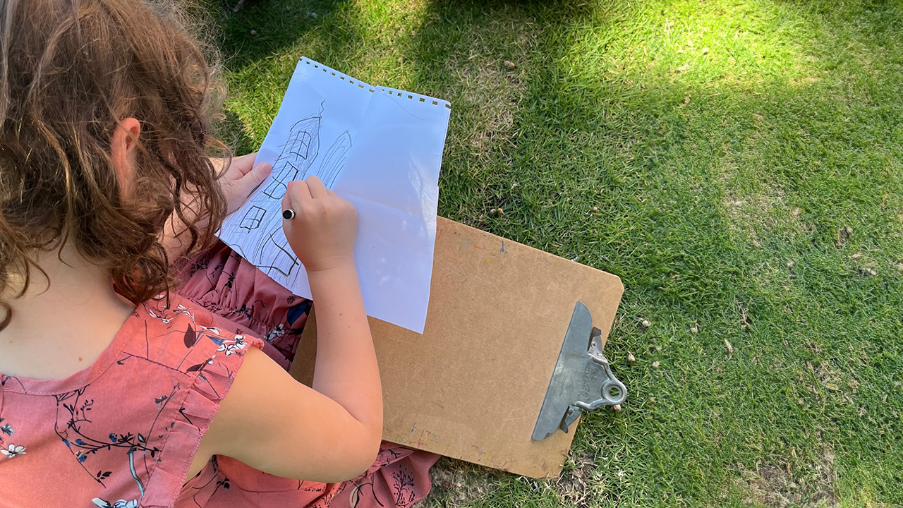 A child sits in the grass drawing a building with pencil and paper