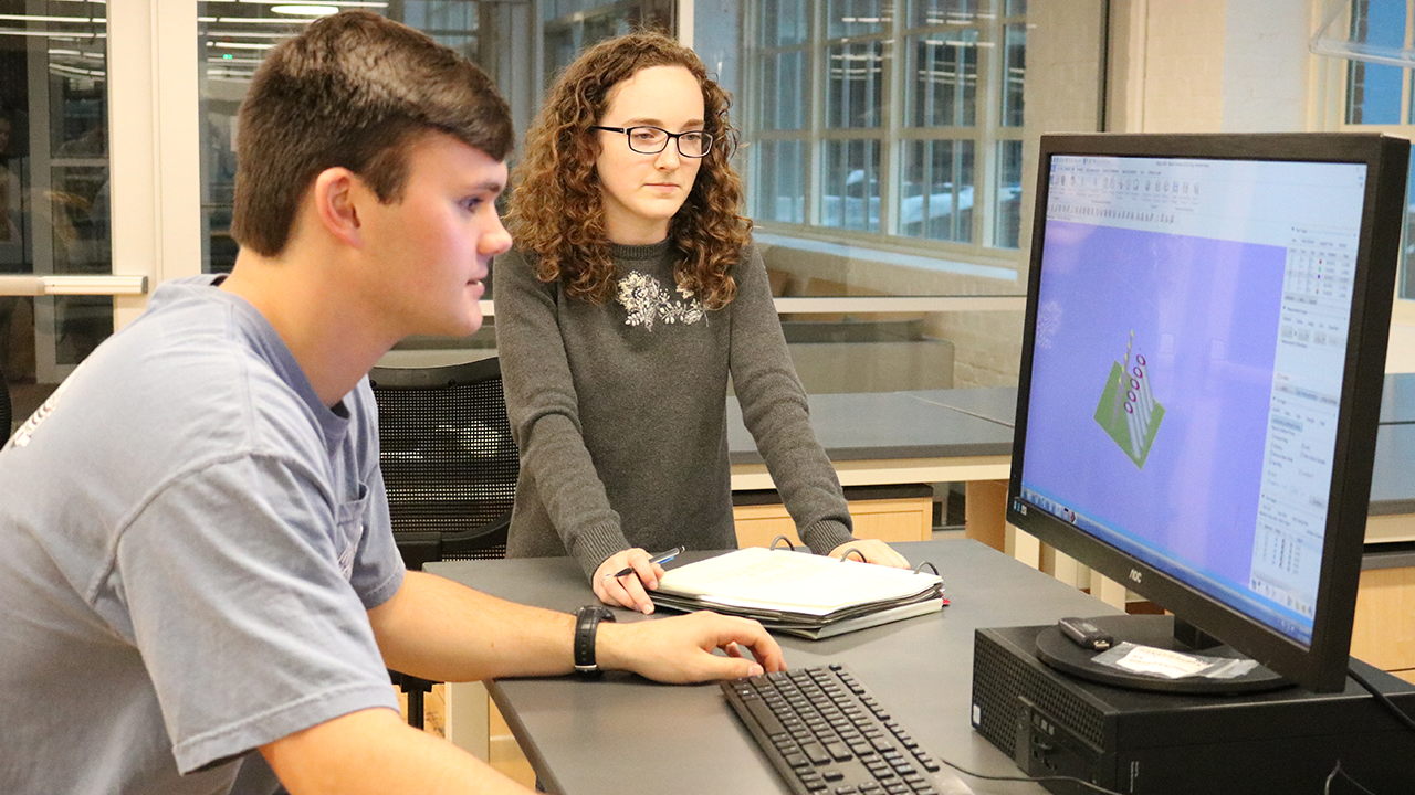 Two students (a male and female presenting) are sitting and standing in front of a large computer looking over data. 