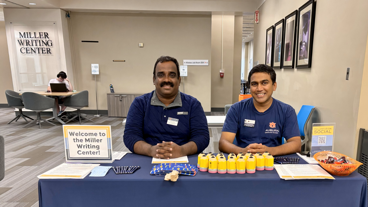 Two students sit at a table ready to give away swag items. A Welcome to the Miller Writing Center sign sits on the table.