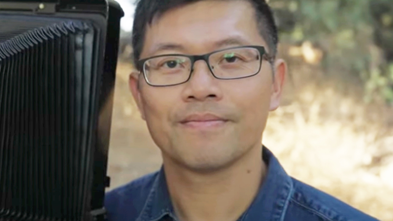 Artist Binh Danh smiles while posing next to his daguerreotype camera. He wears rectangular glasses and a blue denim button-up shirt.