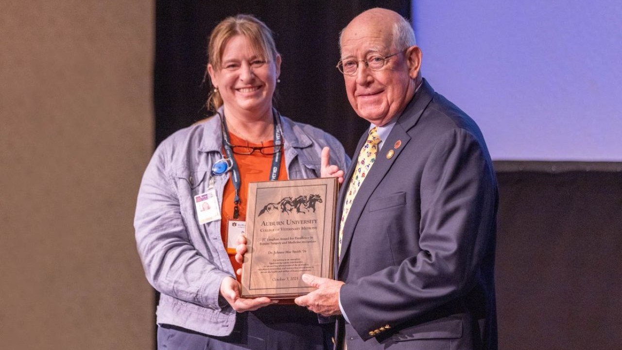 Two individuals standing on a stage holding a wooden plaque with engraved text and horse silhouettes at the top. One person is wearing a suit and tie, and the other is dressed in a casual jacket with a visible badge. A dark backdrop and part of a projection screen are in the background.
