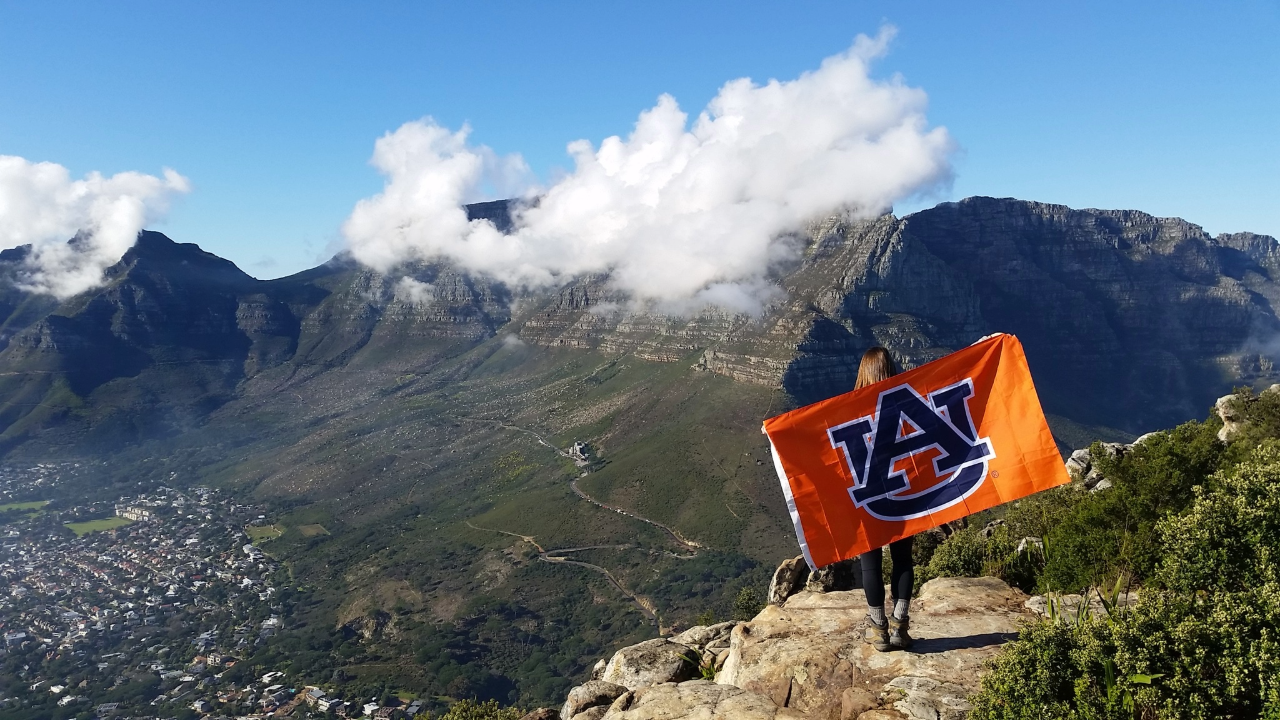 Person stands on a mountain holding an AU flag, More mountains behind them.