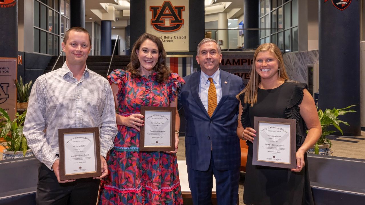 Four individuals standing indoors at an Auburn University venue, each holding a framed award certificate. They are positioned in front of a large Auburn University logo and signage that reads “James and Betty Carroll Atrium.” The setting includes tall columns, plants, and a polished floor, creating a formal presentation environment.