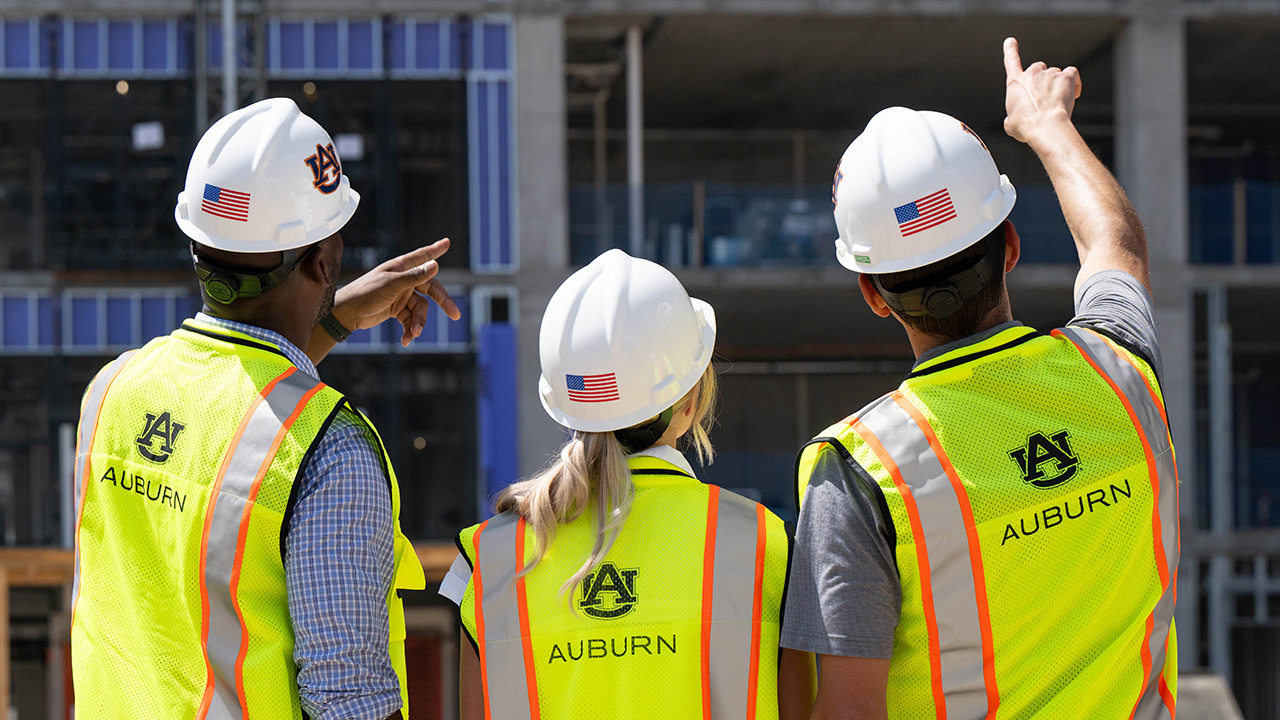 Three people, wearing hard hats and yellow construction vests with an Auburn logo, face away from the camera and point up at a building under construction.