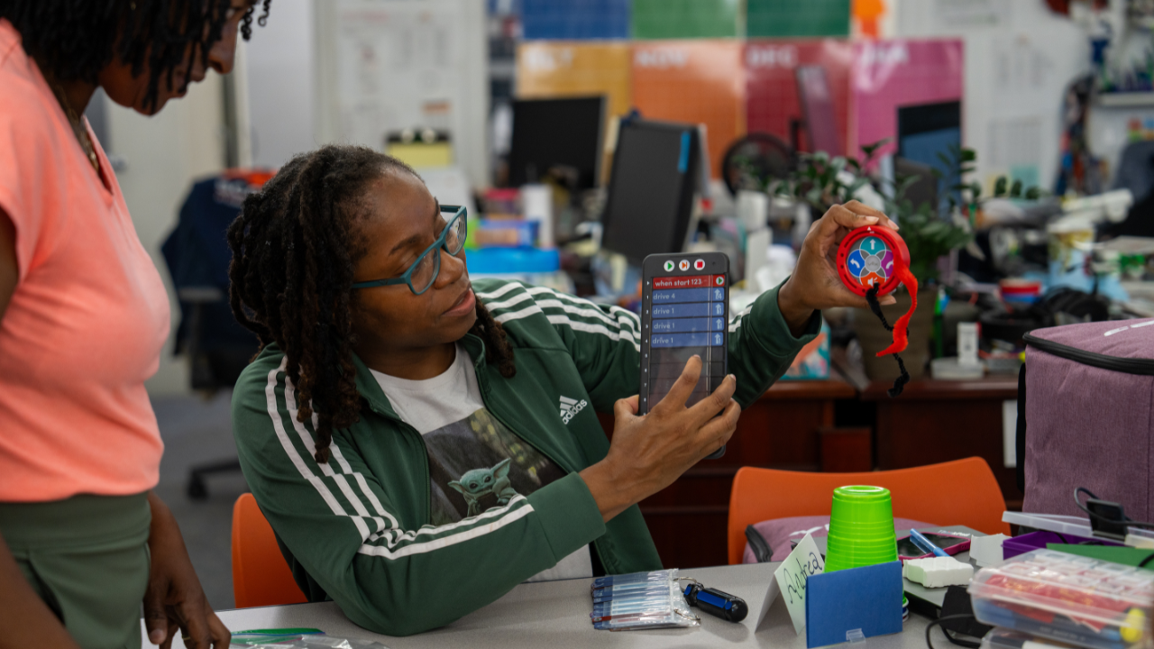 A teacher holds up a VEX 123 robot and a coding tile while speaking with another participant during a computer science training session.