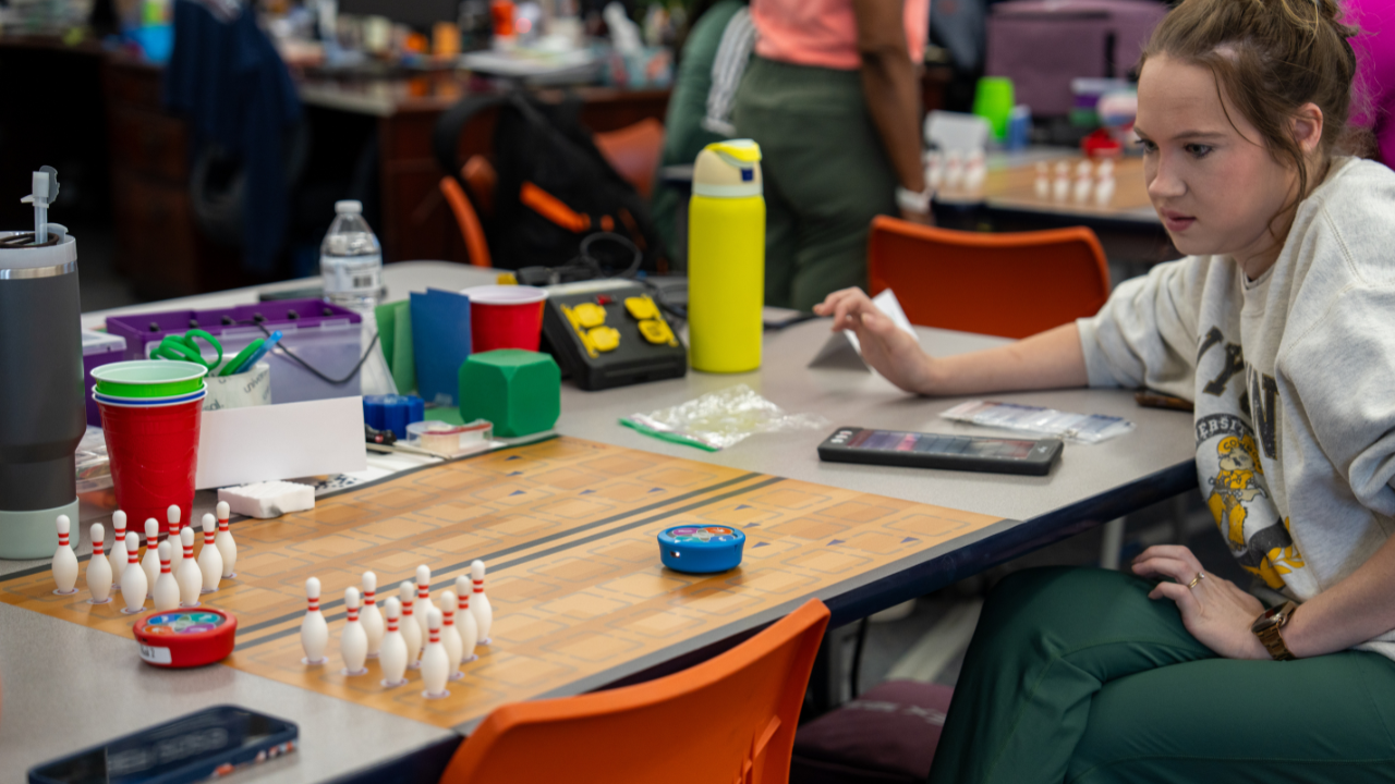 A teacher watches a small blue robot approach 3D-printed bowling pins on a tabletop activity mat