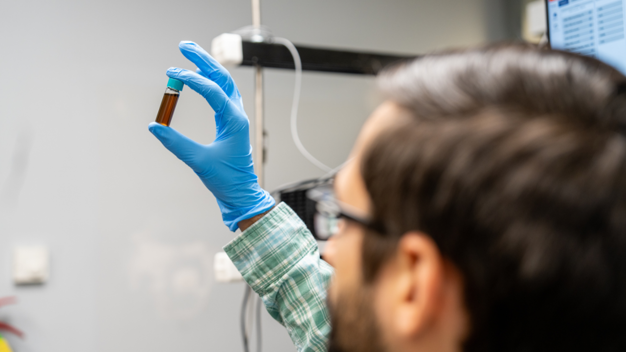 Researcher in safety gloves holding a small vial of amber liquid up to the light in a laboratory setting.