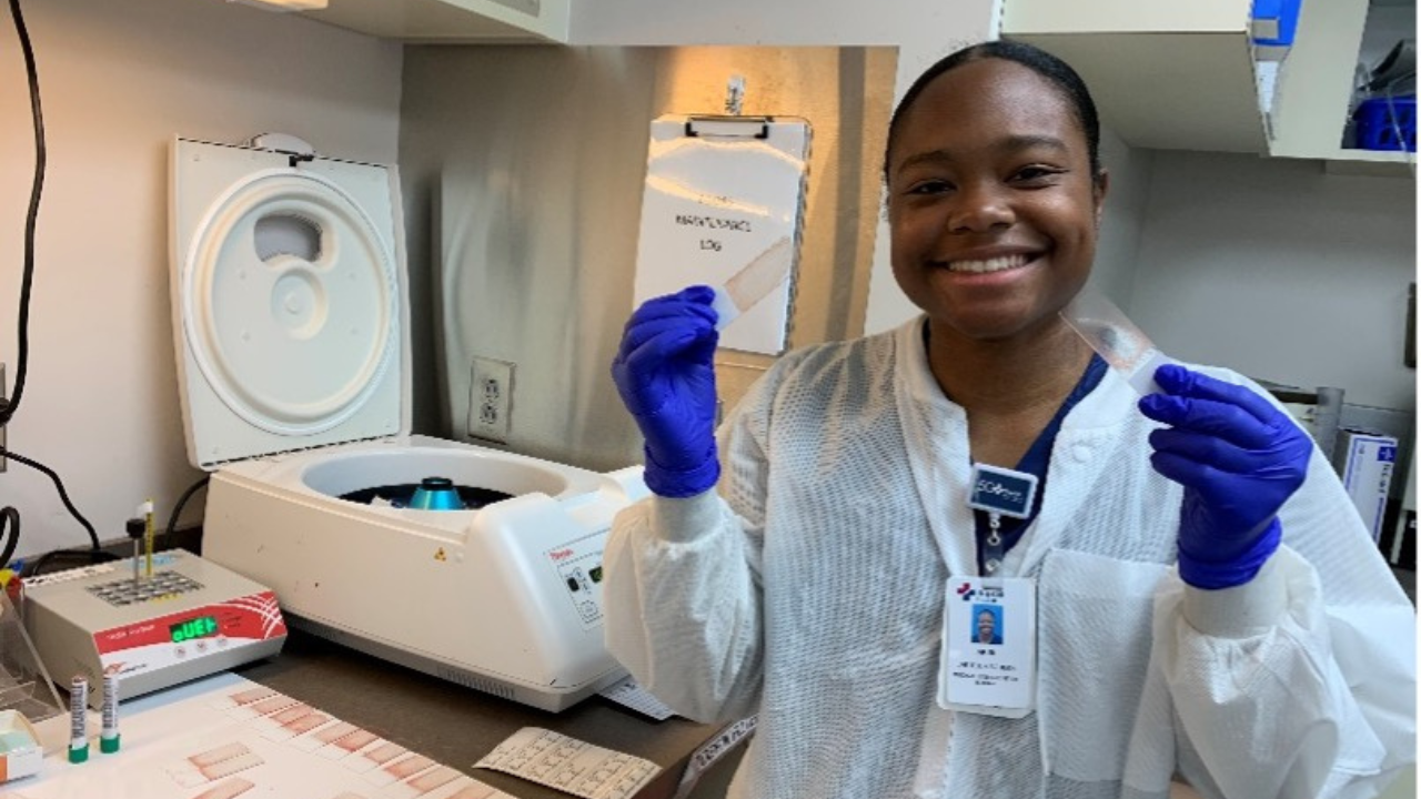 Ariel Sharpe smiles in a medical lab while wearing a white lab coat and purple gloves, holding microscope slides in each hand. A centrifuge and lab equipment are visible on the counter beside her.