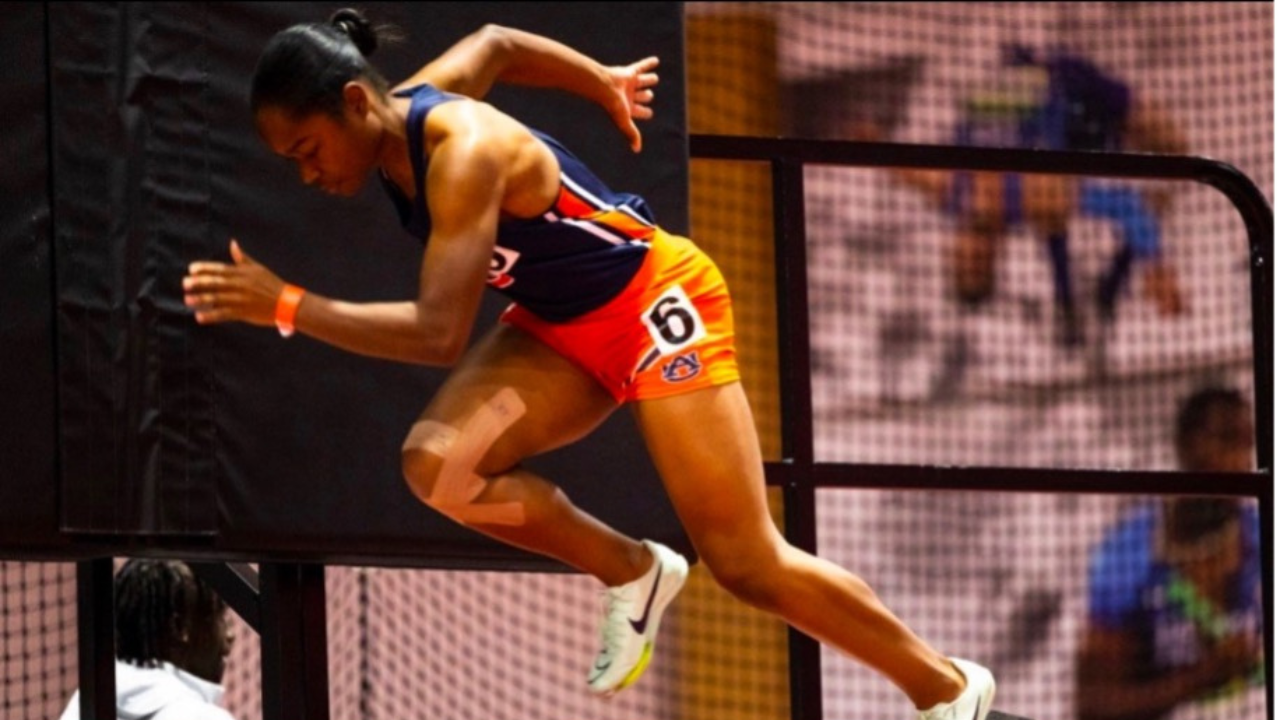 Ariel Sharpe sprints indoors wearing an Auburn track uniform, mid-stride with tape on her knee and determination on her face. A blurred figure watches from behind a netted barrier.