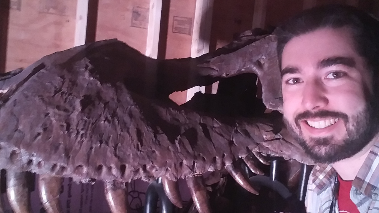 Thomas Cullen smiles next to the fossilized skull of a large predatory dinosaur, its massive teeth visible in the dim light.