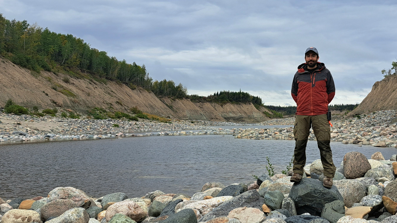 Thomas Cullen stands on rocks at the edge of a river, with steep, tree-lined banks and a cloudy sky in the background.
