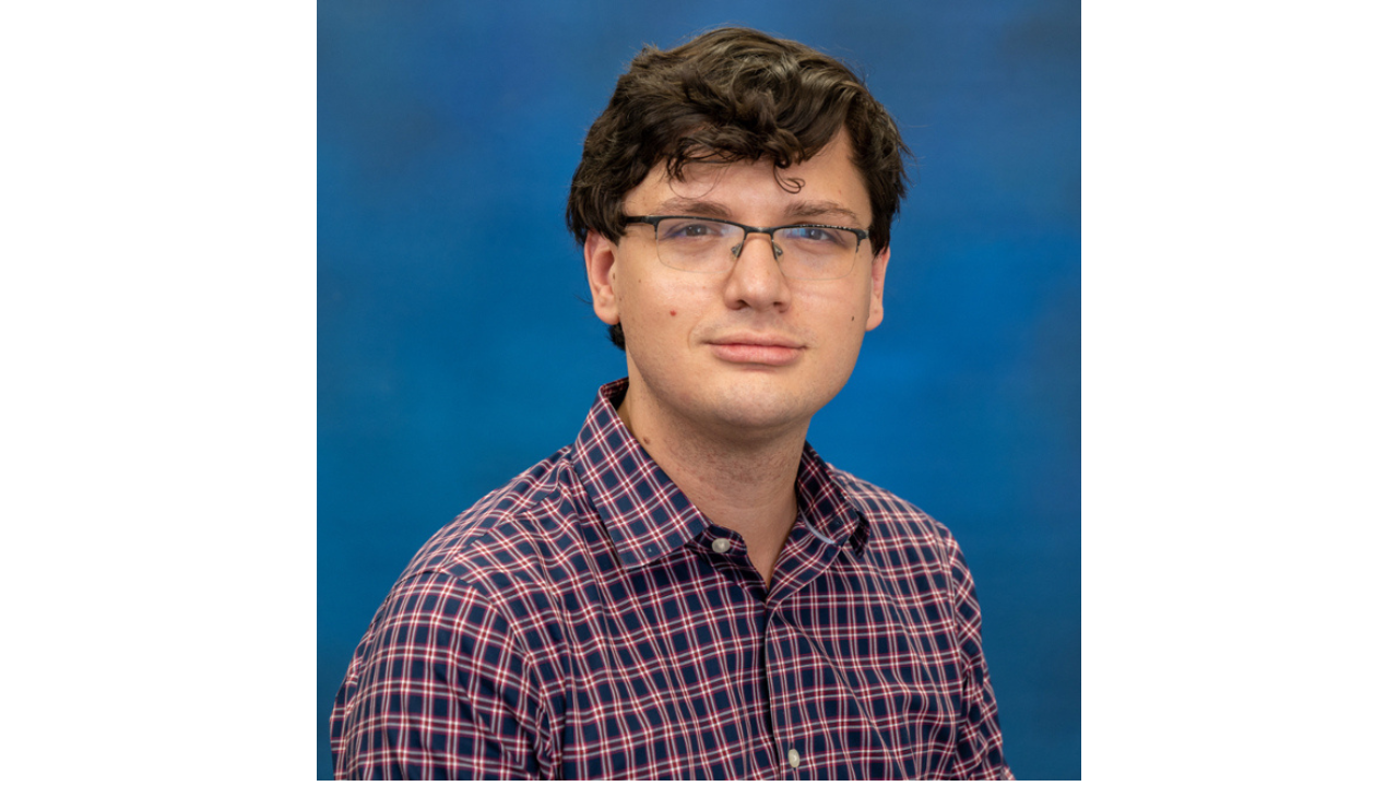 Eric Burkholder poses for a professional portrait in a red and navy plaid shirt against a blue backdrop.