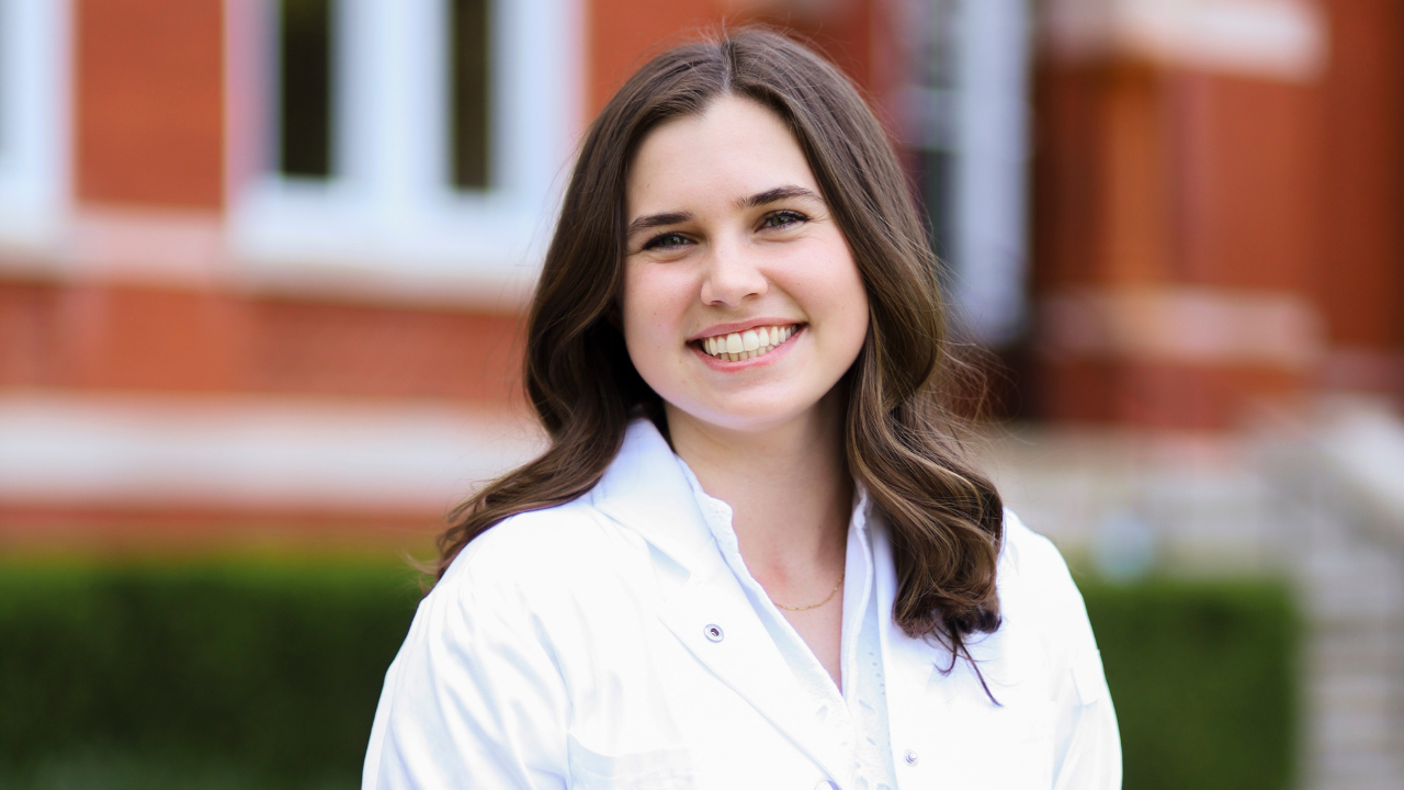 Kaleigh Rice smiles outdoors in front of a red brick building, wearing a white lab coat.