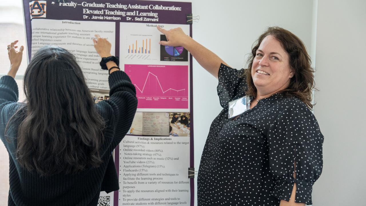 Two women pin up a research poster on a white wall. One faces the poster while the other smiles at the camera.