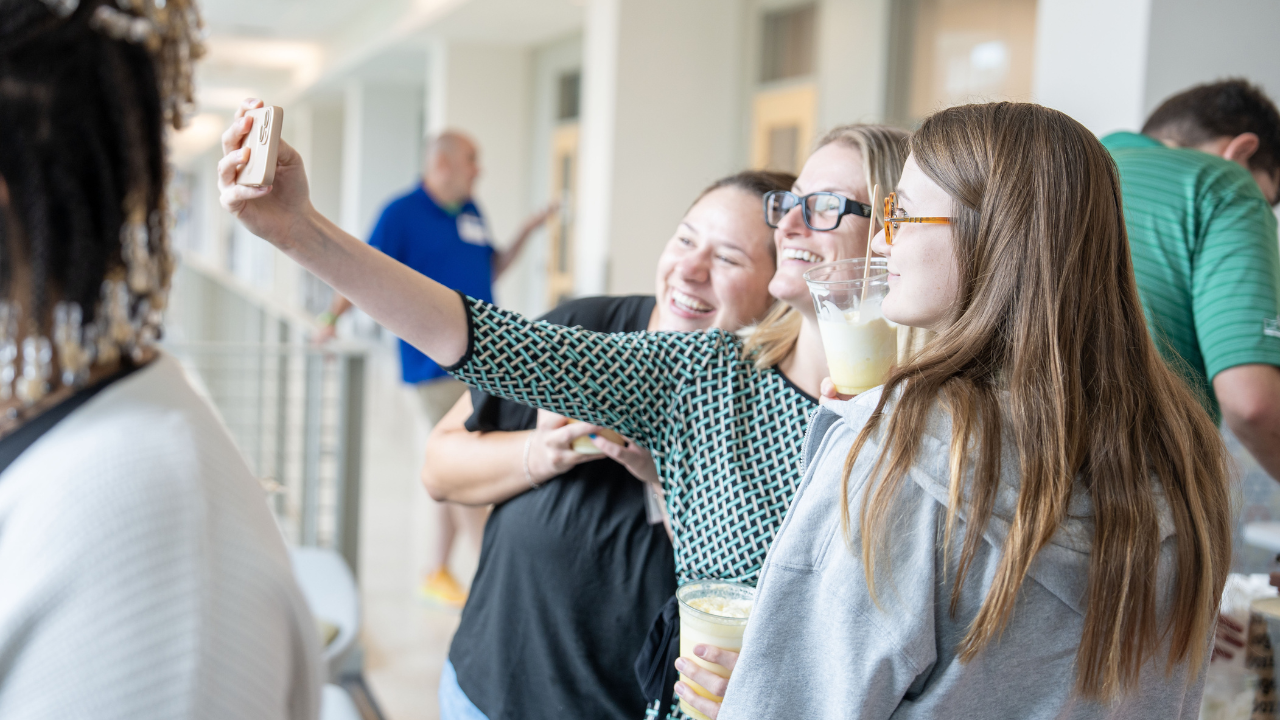 Three women smile and take a selfie together while holding lemonades during an indoor social gathering.