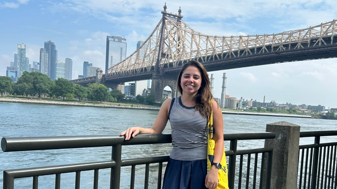 Raissa Santos de Lima Rosa standing by a railing with the Ed Koch Queensboro Bridge and Manhattan skyline in the background.