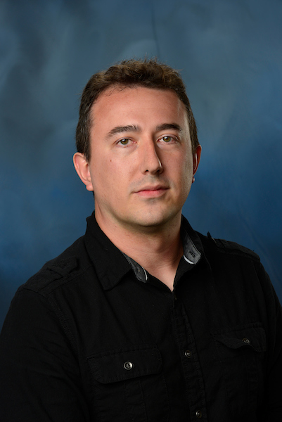 Portrait of Shawn Oset, Auburn University physics graduate student, wearing a black button-up shirt against a blue studio background.