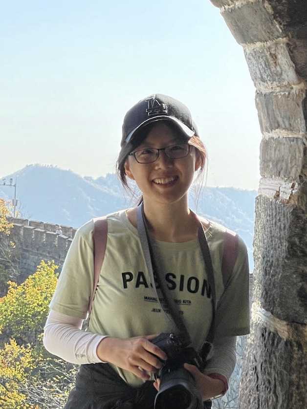 Zexi Xing, Auburn University postdoctoral researcher, smiling while wearing a baseball cap and glasses and holding a camera with mountains in the background.