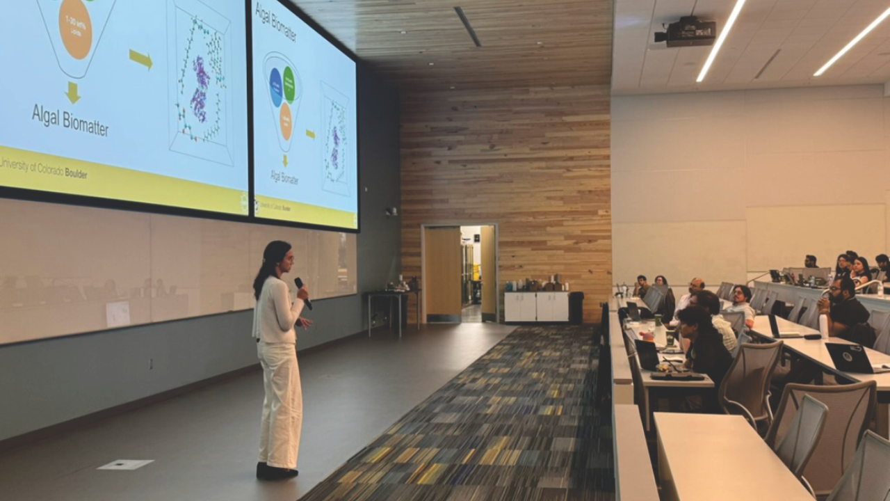 A woman stands at the front of a lecture hall holding a microphone and presenting slides about algal biomatter to an audience seated with laptops.
