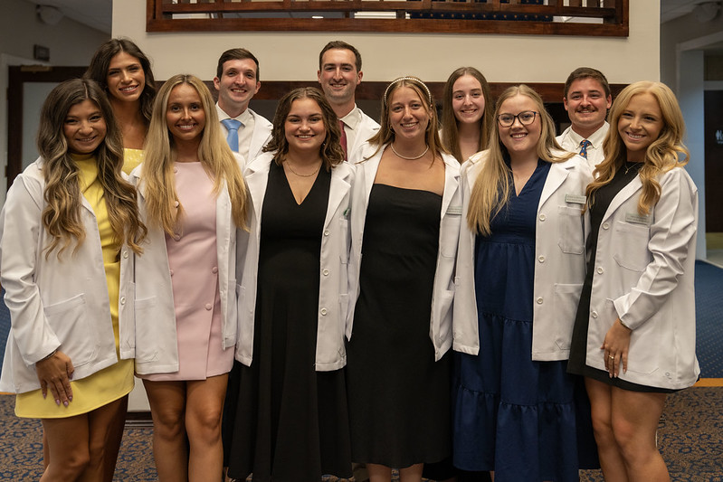 A group of eleven Rural Medicine Program students wearing white coats stand together, smiling for a photo inside the ceremony venue.