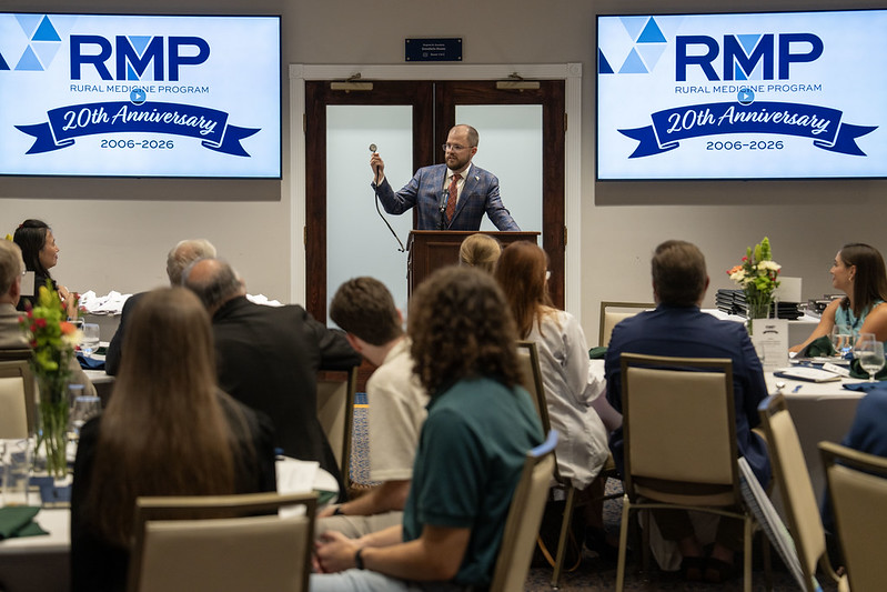 A man in a suit, Dr. Tim Littman, stands at a podium holding up a stethoscope while speaking to a seated audience at the Rural Medicine Program’s 20th anniversary ceremony.