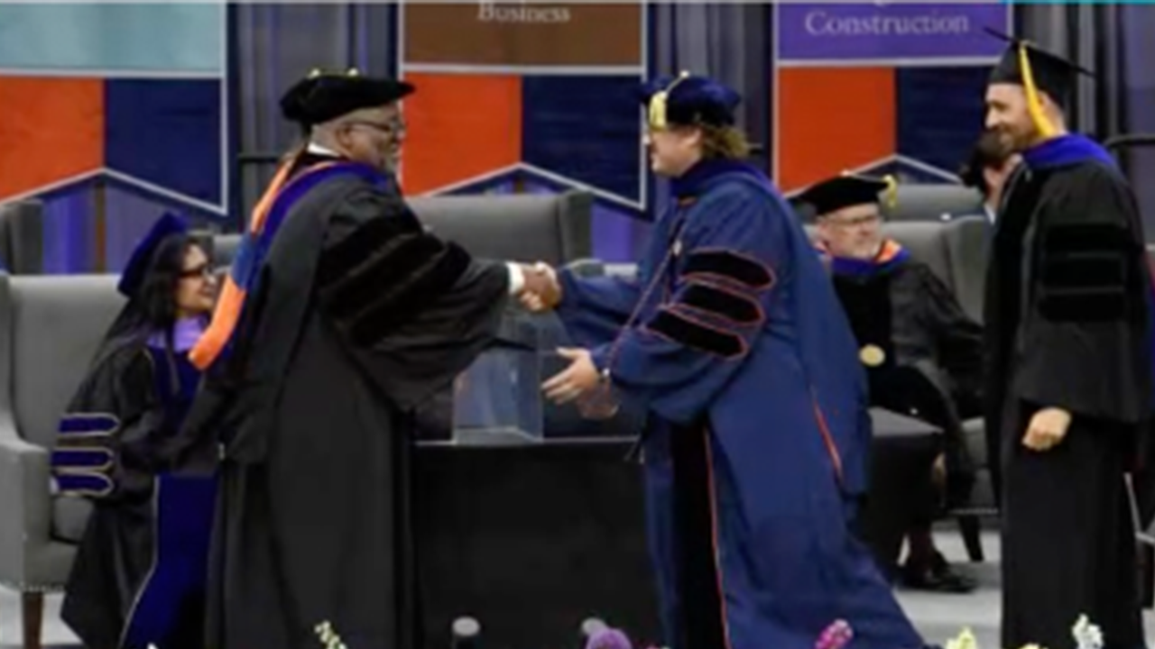 A doctoral graduate in full regalia shakes hands with a faculty member on stage during a commencement ceremony. Several other faculty members in regalia are seated in the background.