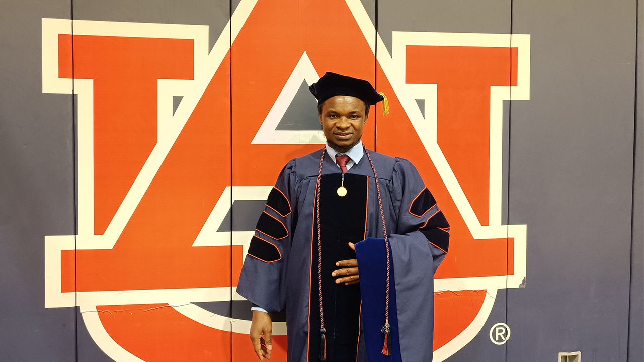 A graduate in doctoral regalia stands proudly in front of a large Auburn University logo wall. He wears a navy gown with black velvet panels, orange piping, a tam with a gold tassel, and a medallion.