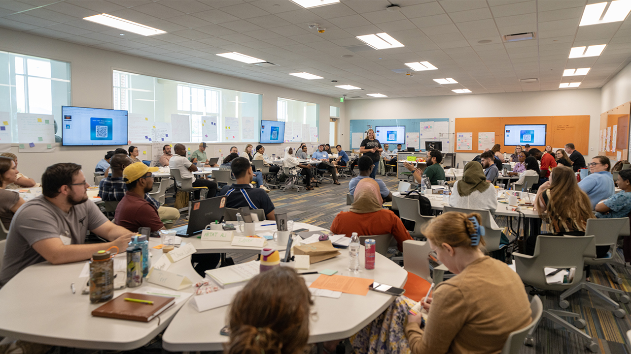 Wide view of a classroom filled with participants working in groups and listening to a facilitator at the Summer STEM Institute