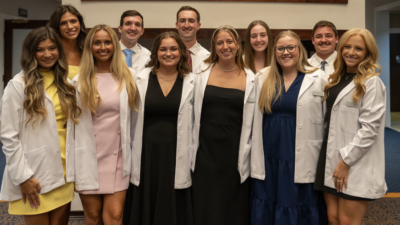 A group of eleven Rural Medicine Program students wearing white coats stand together, smiling for a photo inside the ceremony venue.