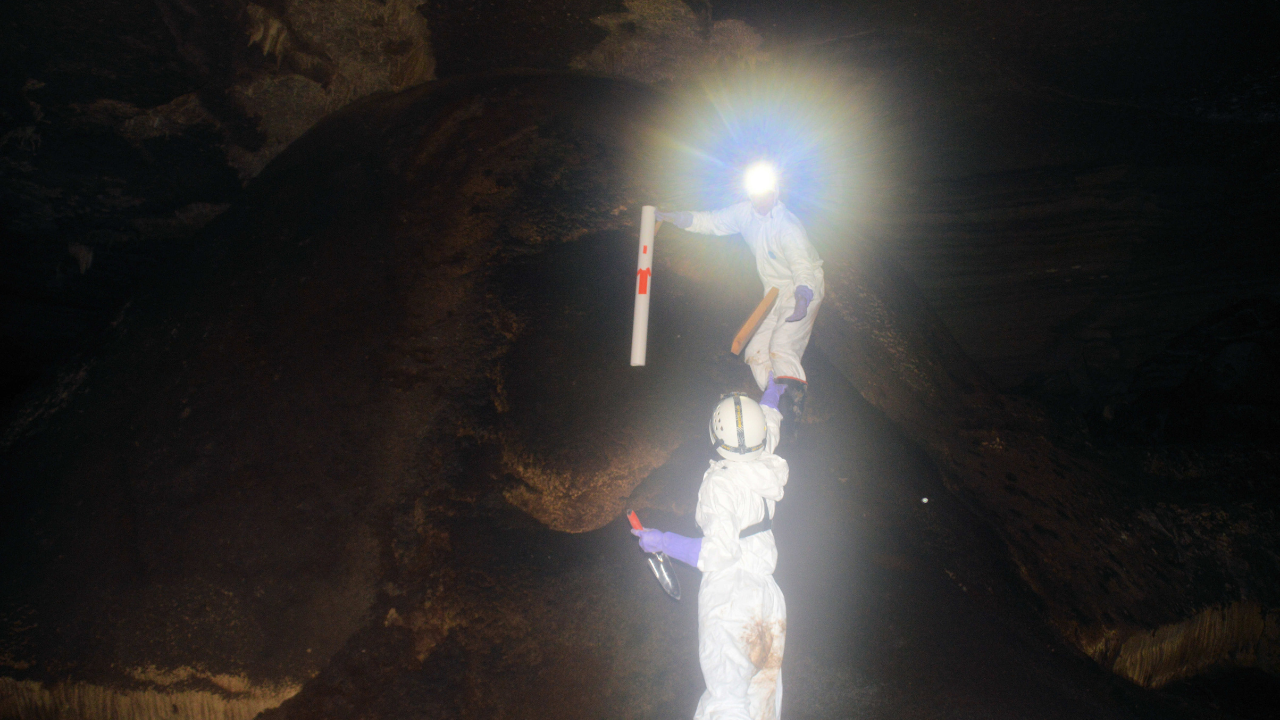 Two researchers wearing helmets and white protective suits work together inside a cave, passing a cylindrical core tube.