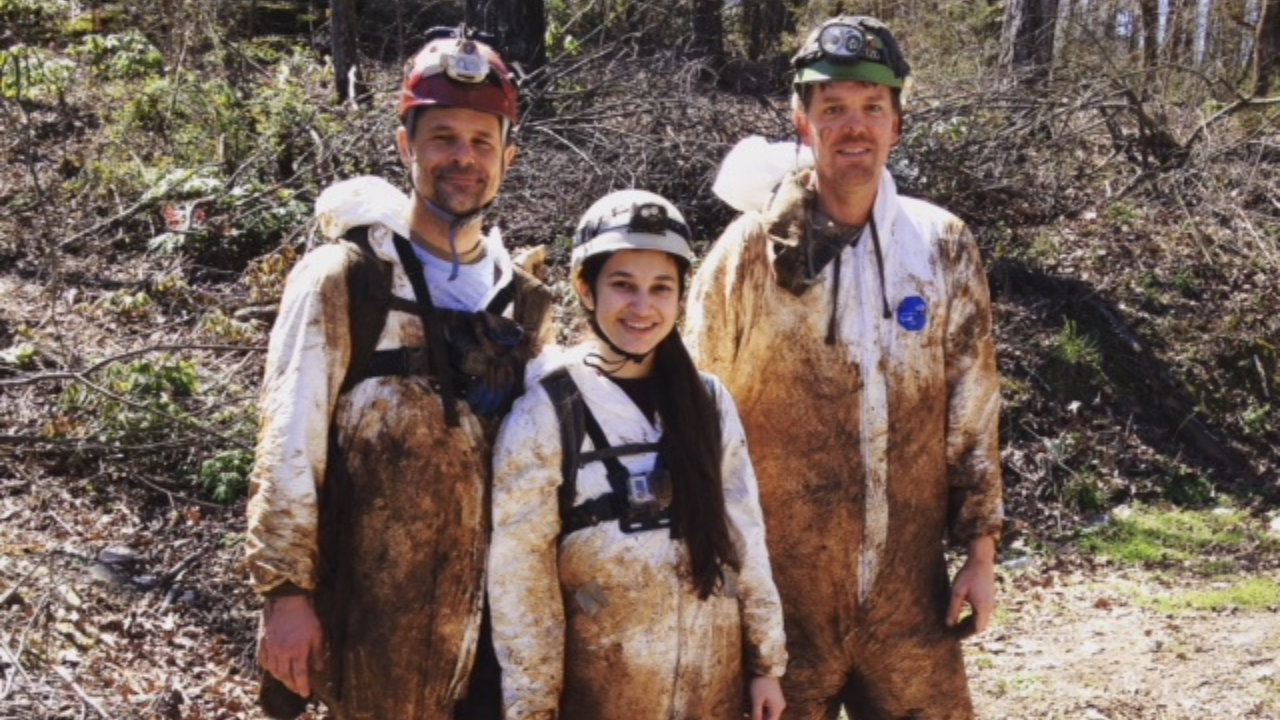 Three researchers wearing mud-covered protective suits and helmets stand outdoors after a cave expedition.