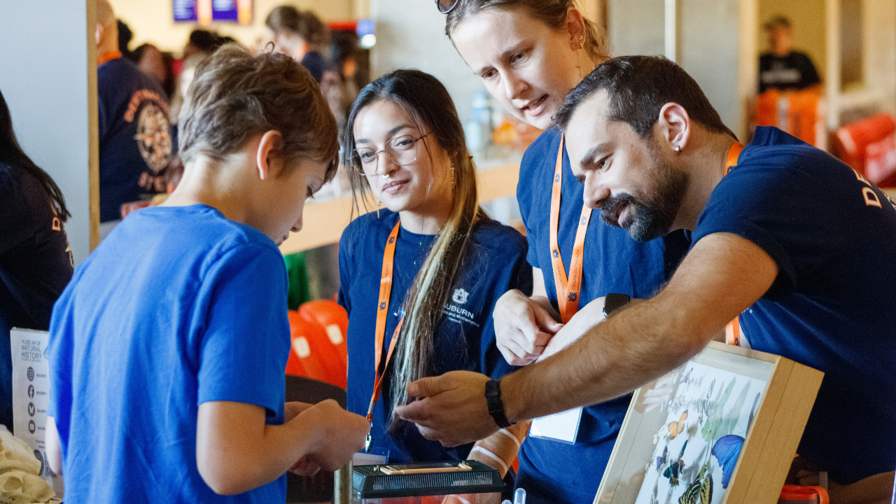 A COSAM volunteer explains a science exhibit to a student as two other volunteers look on.