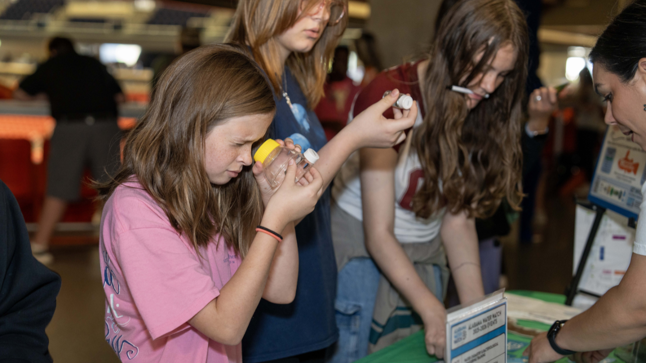 A student peers closely through a clear plastic container while examining a sample at a biology exhibit table.