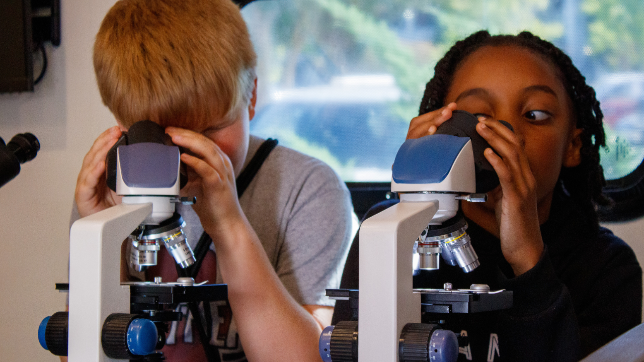 Two students look through microscopes at samples during a mobile science lab activity.
