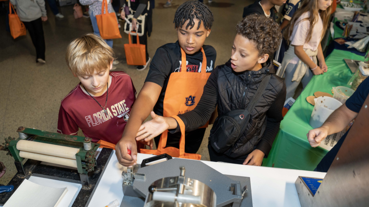 Three middle school students explore a hands-on STEM exhibit using a machine with rollers at Auburn University’s Destination STEM Expo.