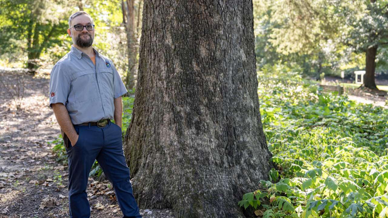 Patrick Thompson, wearing glasses and a gray Auburn shirt, stands beside the wide trunk of a large oak tree surrounded by undergrowth.