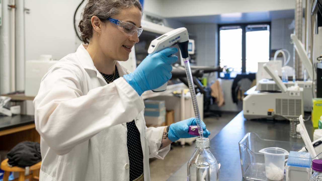 Katie Rush, in a white lab coat and blue gloves, uses a pipette to transfer liquid into a glass bottle in her Auburn University biochemistry lab. Laboratory instruments and supplies are visible in the background.