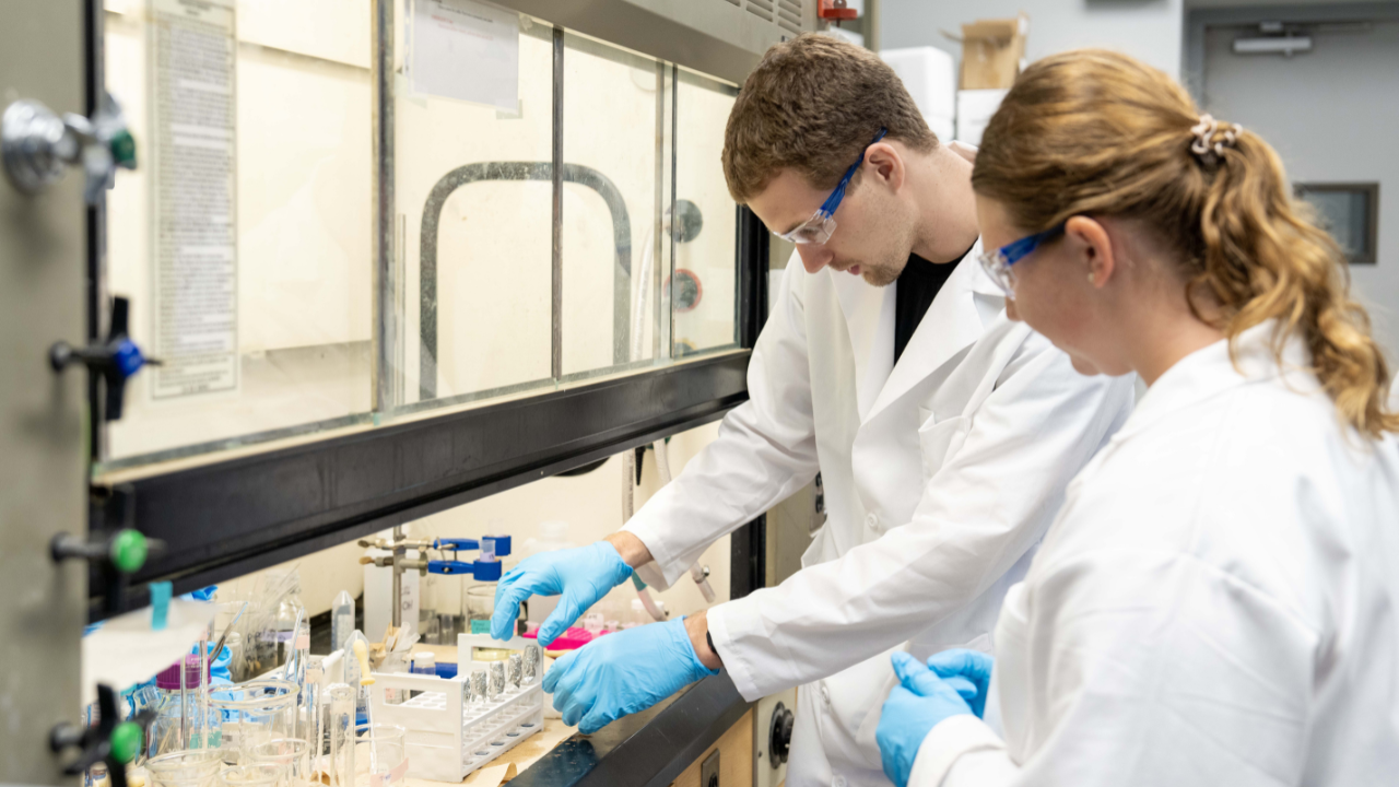 Two graduate students wearing lab coats, gloves, and safety glasses work together at a lab bench, measuring samples in test tubes inside a chemical fume hood.