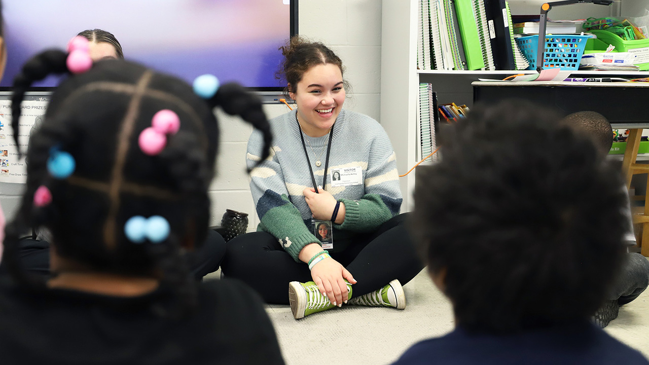 A young woman sits on the floor smiling at a group of students