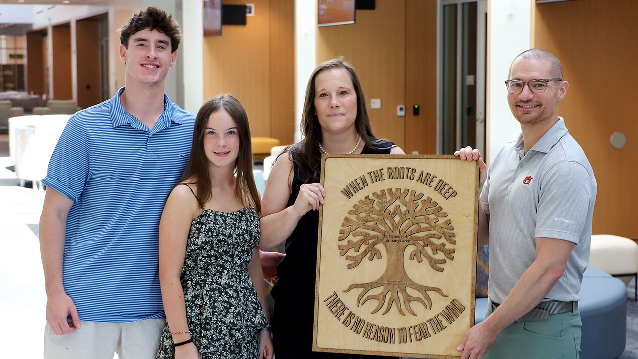 Dr. Andrew Pendola is pictured inside the new College of Education building along with the family of Brittany Larkin. Pendola is holding a wooden memorial in honor of Dr. Larkin.