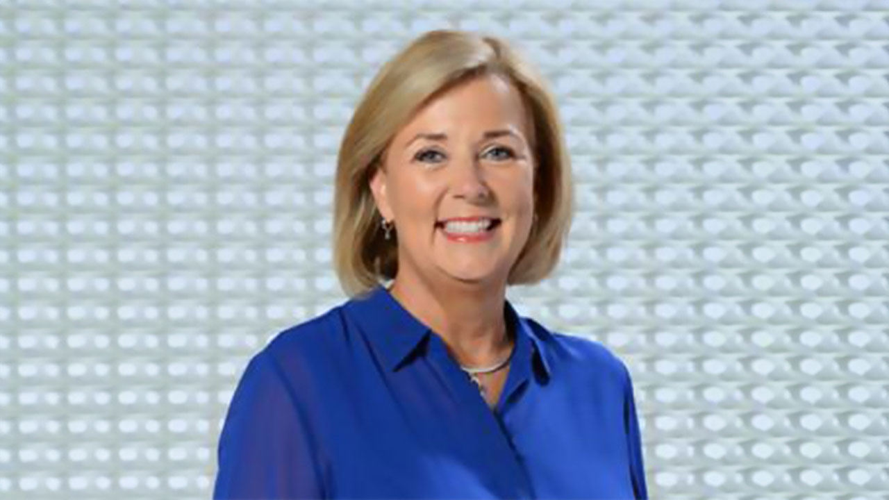 Sue Steele smiles in a posed photo with a light blue patterned design behind her.