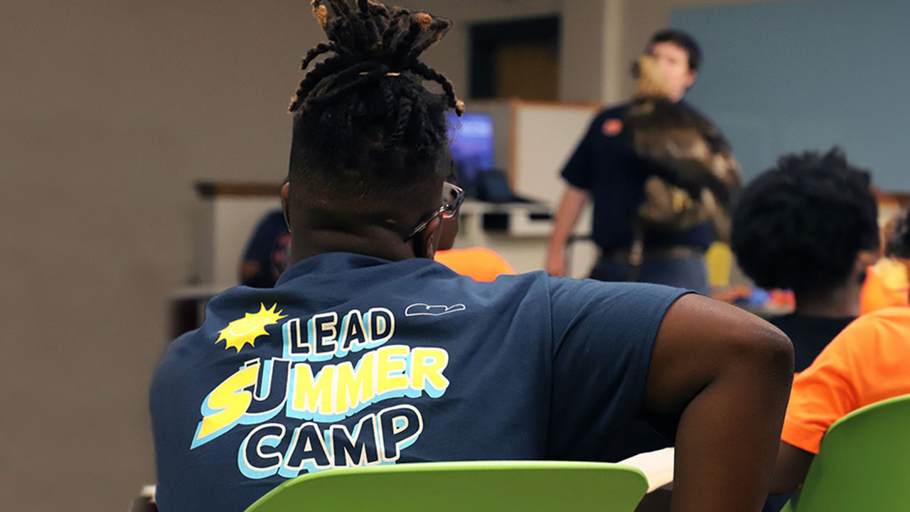 A camper in the Learners Exploring Academic Dreams initiative is pictured sitting in a clasroom, with his back to the camera and wearning a shirt with the camp logo displayed.