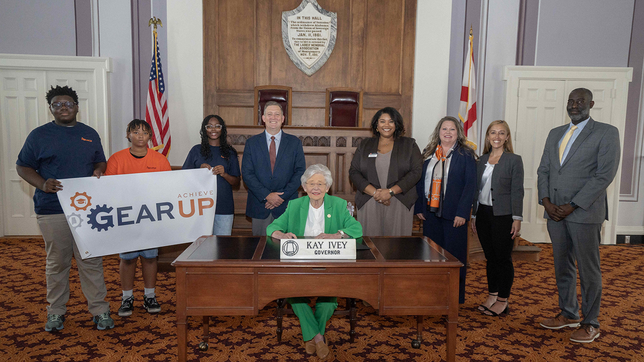 A delegation of students and staff from GEAR UP Achieve stand on either side of Alabama Governor KAy Ivey, who is seated at a desk.